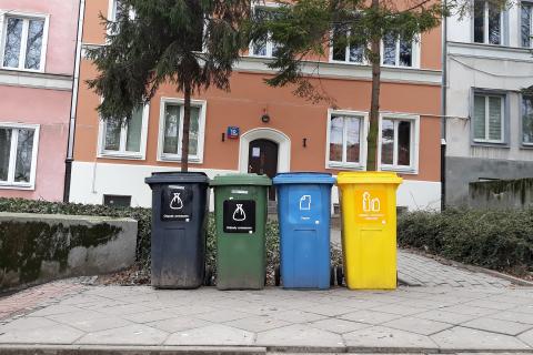 Cztery kubły w różnych kolorach przeznaczone na poszczególne frakcje odpadów stoją na krawędzi chodnika przed kamienicą